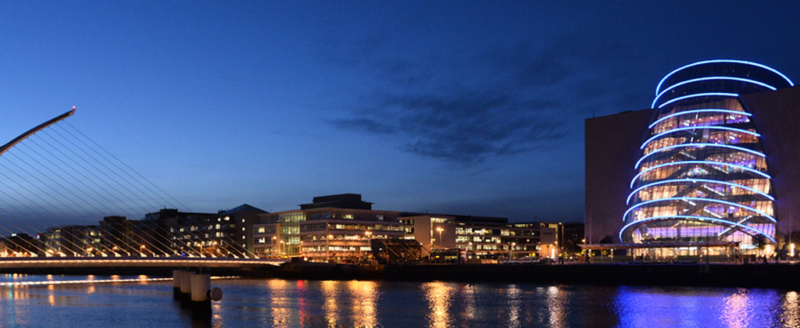 The brightly lit early evening cityscape of downtown Dublin, Ireland, is presented. A body of water is visible in the forefront and a suspension bridge can be seen on the left-hand side of the image.